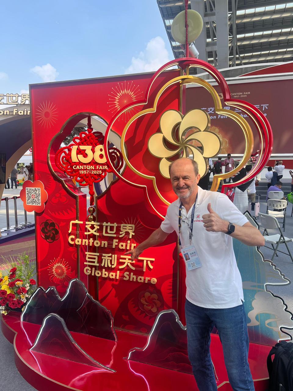 Homem sorridente posando em frente ao grande display vermelho e dourado de boas-vindas da 138ª Canton Fair em Guangzhou, com os dizeres 'Global Share' e as datas 1957-2025.